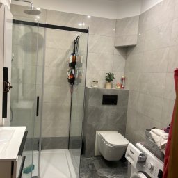 KONRAD KRAUZE - Nowoczesna łazienka po remoncie: szare płytki imitujące marmur, kabina prysznicowa z czarnymi profilami, podwieszana toaleta i pralka. Minimalistyczny design.