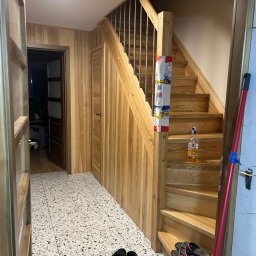 KONRAD KRAUZE - Korytarz po remoncie: drewniane schody z metalową barierką, drewniana boazeria na ścianach, wzorzysta podłoga. Widok z perspektywy wejścia, z otwartymi drzwiami.