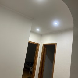 KONRAD KRAUZE - Białe wnętrze z dwiema otwartymi, drewnianymi ościeżnicami drzwiowymi, widok na podłogę i sufit z oświetleniem punktowym, łukowate przejście.