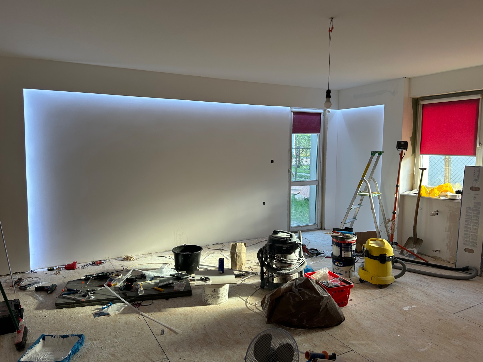 Wnętrze w trakcie remontu: białe ściany z podświetleniem LED, narzędzia malarskie, drabina, okno z czerwoną roletą. Widoczny bałagan remontowy na podłodze.