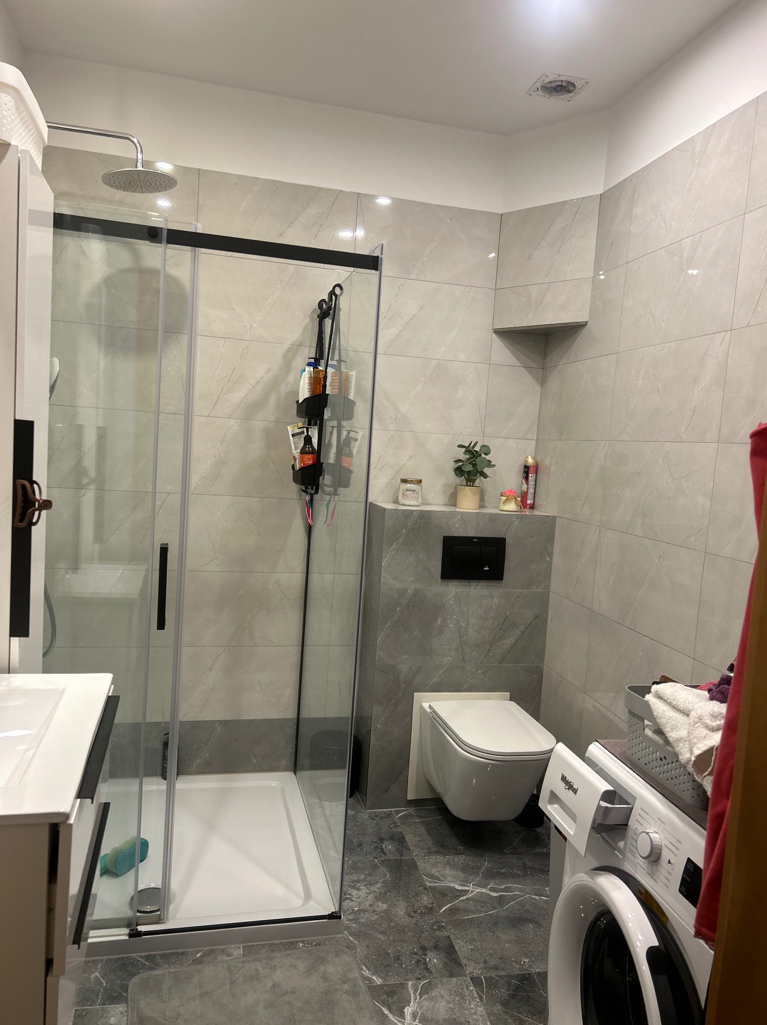 Nowoczesna łazienka po remoncie: szare płytki imitujące marmur, kabina prysznicowa z czarnymi profilami, podwieszana toaleta i pralka. Minimalistyczny design.
