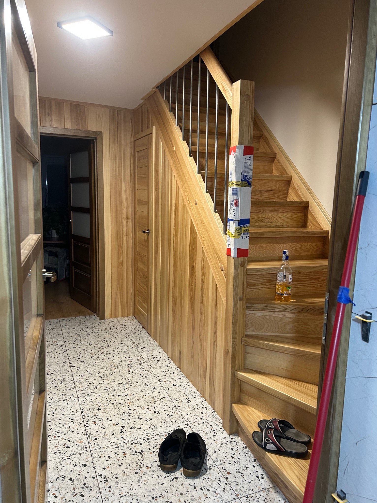 Korytarz po remoncie: drewniane schody z metalową barierką, drewniana boazeria na ścianach, wzorzysta podłoga. Widok z perspektywy wejścia, z otwartymi drzwiami.