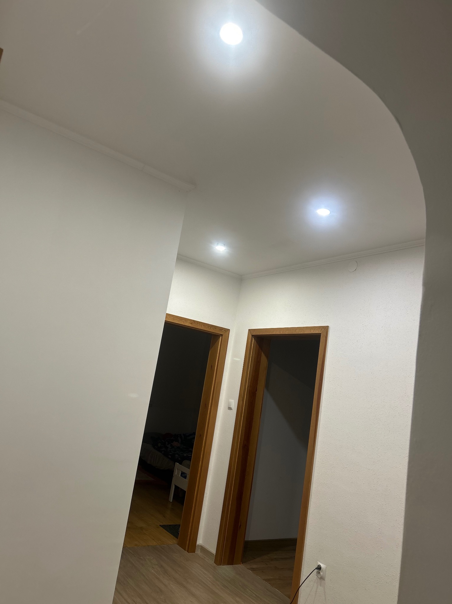 Białe wnętrze z dwiema otwartymi, drewnianymi ościeżnicami drzwiowymi, widok na podłogę i sufit z oświetleniem punktowym, łukowate przejście.