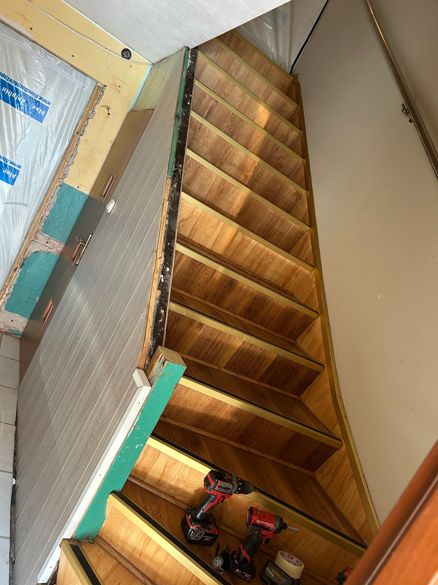 Korytarz w trakcie remontu: widoczne schody z drewnopodobnymi panelami, częściowo wykończona ściana z panelami, narzędzia budowlane na podłodze. Ujęcie z dołu.