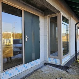 Justyna Banaśkiewicz-Wesołowska Przedsiębiorstwo Handlowo-Usługowe WEBA - Nowe okna i drzwi z szarymi ramami zamontowane w surowym budynku z białego betonu komórkowego. Widoczne pianki montażowe i folia ochronna na dole.