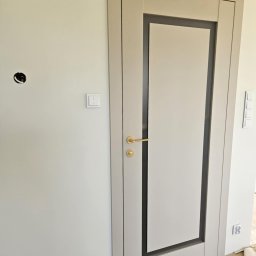 Justyna Banaśkiewicz-Wesołowska Przedsiębiorstwo Handlowo-Usługowe WEBA - Jasne, nowoczesne drzwi wewnętrzne z szarą ramą i złotą klamką, zamontowane w ścianie. Widoczny włącznik światła i otwór na gniazdko elektryczne.