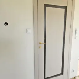 Jasne, nowoczesne drzwi wewnętrzne z szarą ramą i złotą klamką, zamontowane w ścianie. Widoczny włącznik światła i otwór na gniazdko elektryczne.