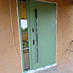 Justyna Banaśkiewicz-Wesołowska Przedsiębiorstwo Handlowo-Usługowe WEBA - Świeżo zamontowane, zielone drzwi zewnętrzne z długim, czarnym uchwytem, z szybą boczną. Uszczelnione pianką montażową i taśmą ochronną Veka.