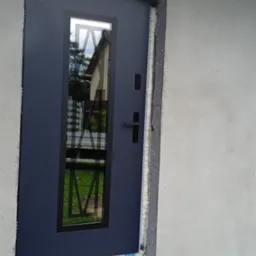 Ciemne, nowoczesne drzwi zewnętrzne z szybą, zamontowane w surowym otworze, widoczne pianki montażowe i folia ochronna VEKA, częściowo otynkowana ściana.