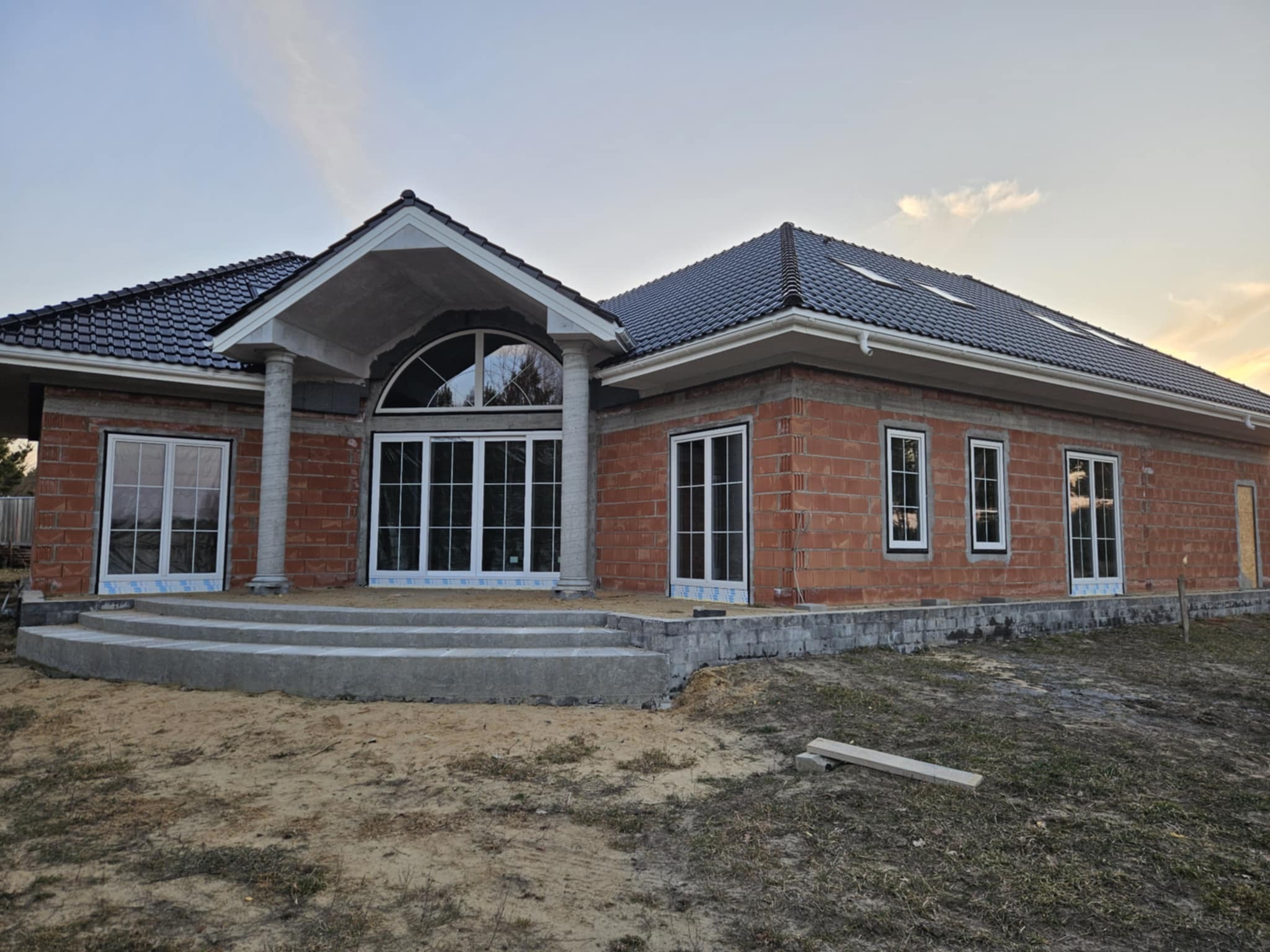 Dom w trakcie budowy z białymi oknami PCV i dużym oknem łukowym. Widoczne cegły i betonowe schody wejściowe. Dach z ciemnej dachówki.