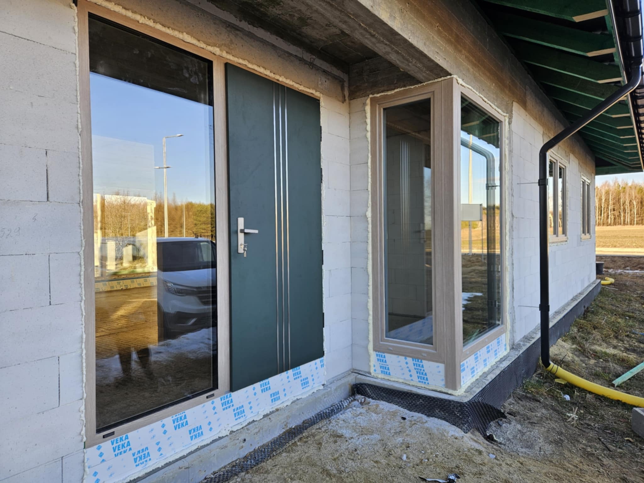 Nowe okna i drzwi z szarymi ramami zamontowane w surowym budynku z białego betonu komórkowego. Widoczne pianki montażowe i folia ochronna na dole.