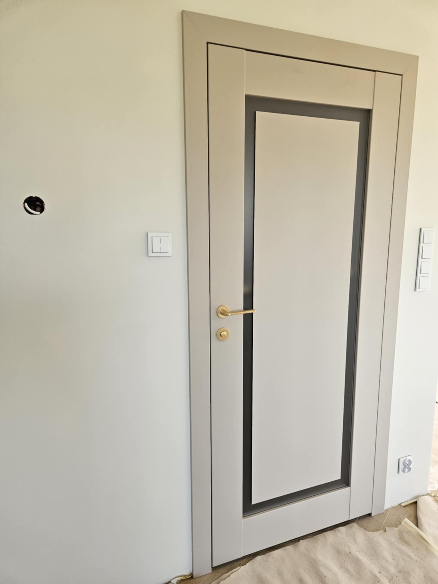 Jasne, nowoczesne drzwi wewnętrzne z szarą ramą i złotą klamką, zamontowane w ścianie. Widoczny włącznik światła i otwór na gniazdko elektryczne.