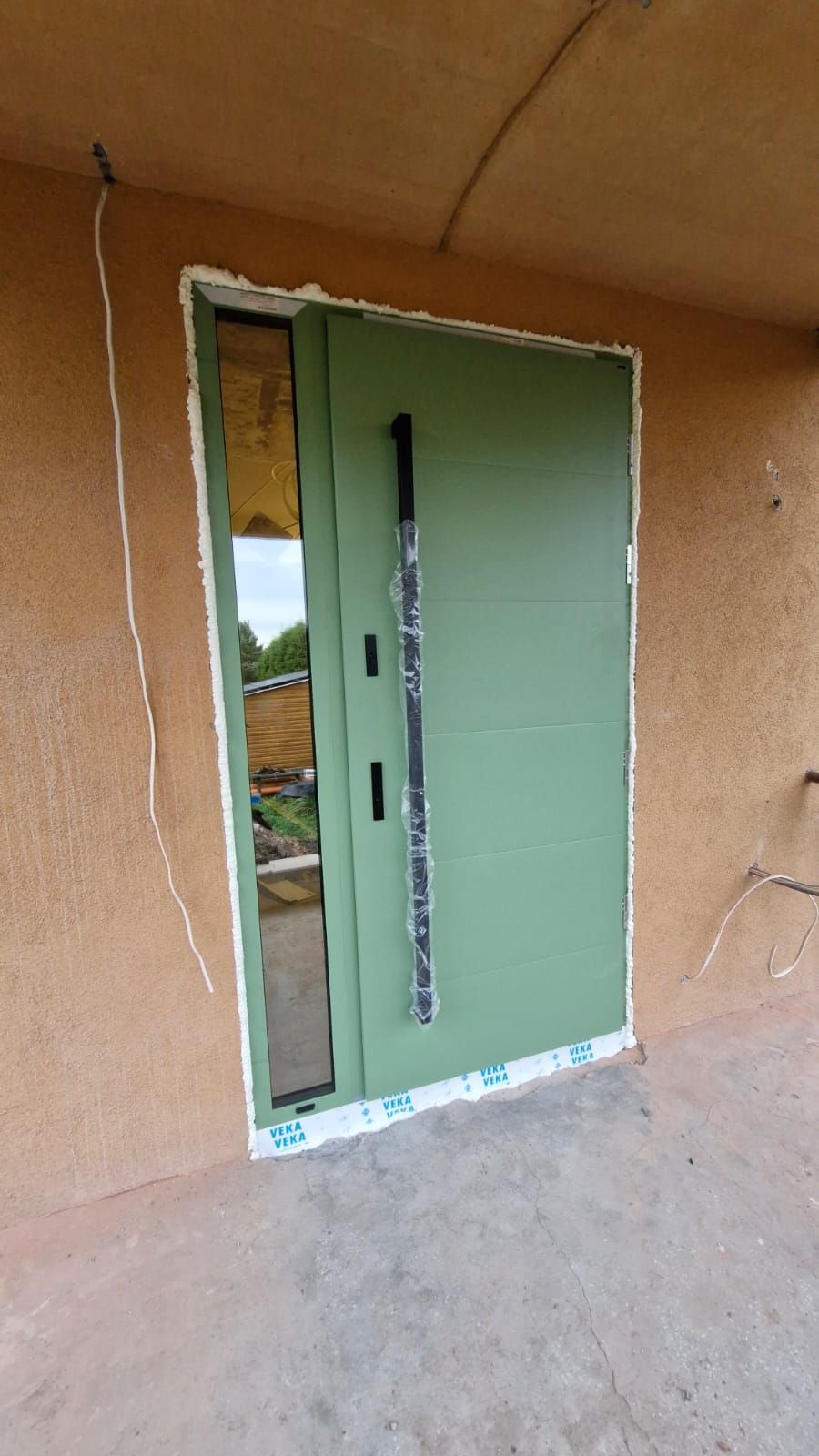 Świeżo zamontowane, zielone drzwi zewnętrzne z długim, czarnym uchwytem, z szybą boczną. Uszczelnione pianką montażową i taśmą ochronną Veka.