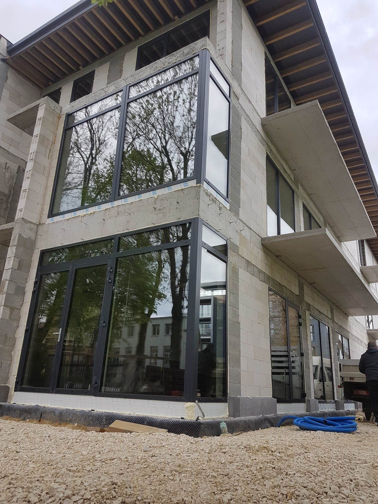 Nowoczesny budynek w trakcie budowy z dużymi, czarnymi oknami. Widoczne surowe ściany z betonu komórkowego i odbicie drzew w szybach.