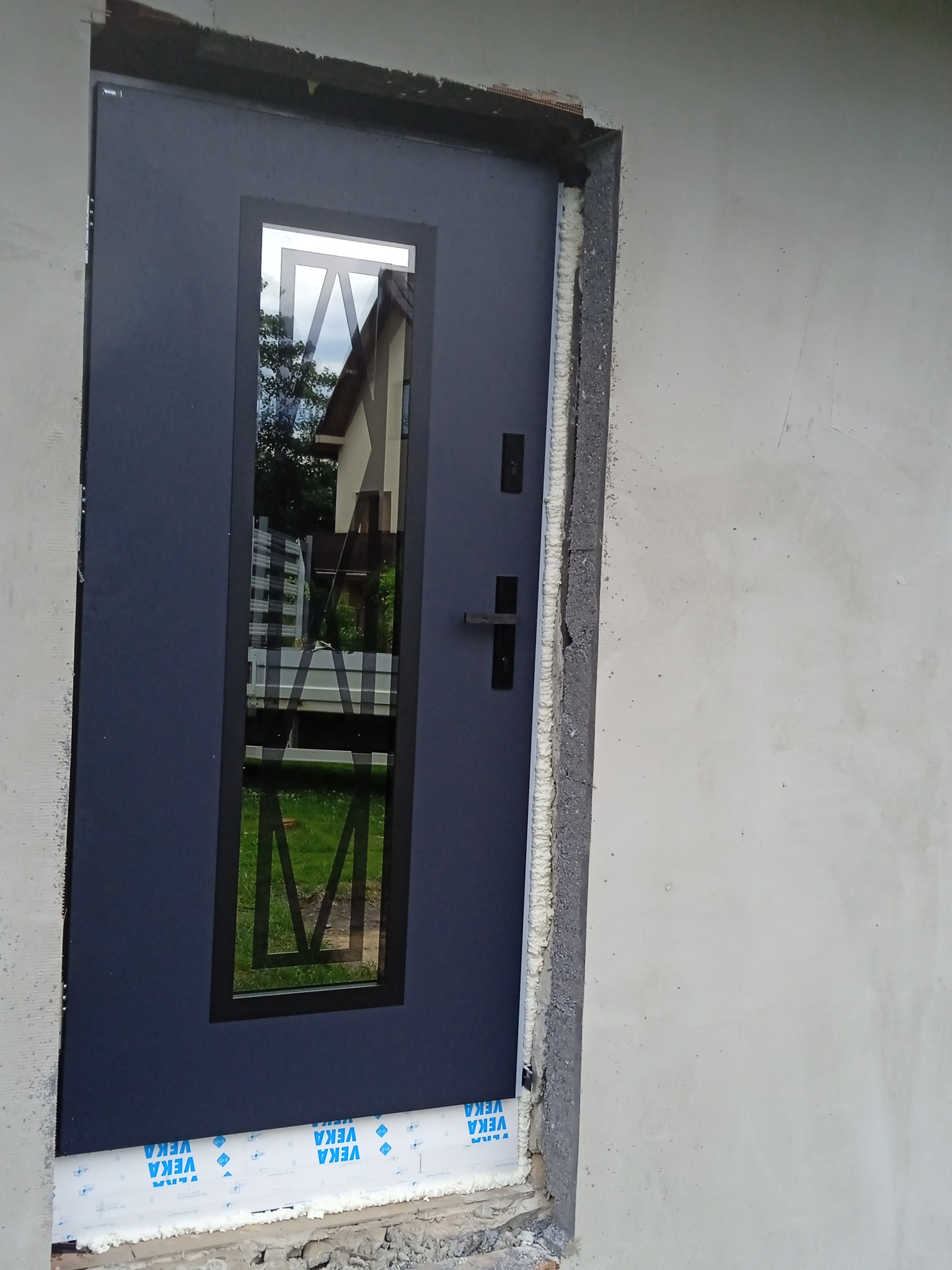 Ciemne, nowoczesne drzwi zewnętrzne z szybą, zamontowane w surowym otworze, widoczne pianki montażowe i folia ochronna VEKA, częściowo otynkowana ściana.