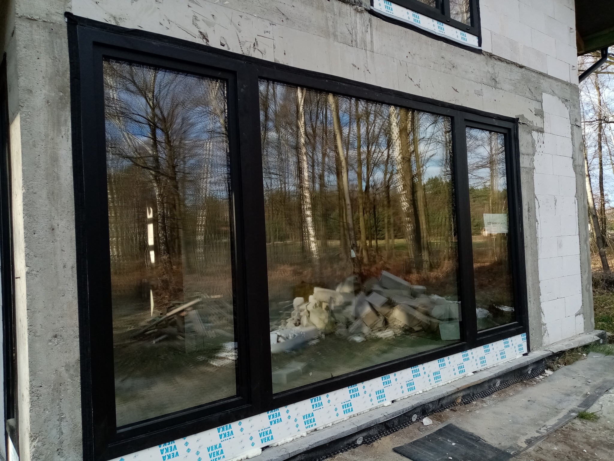 Zamontowane okno z czarną ramą w surowym stanie budynku, widoczna izolacja i gruz wewnątrz. W szybach odbija się las. Detal montażu okna.