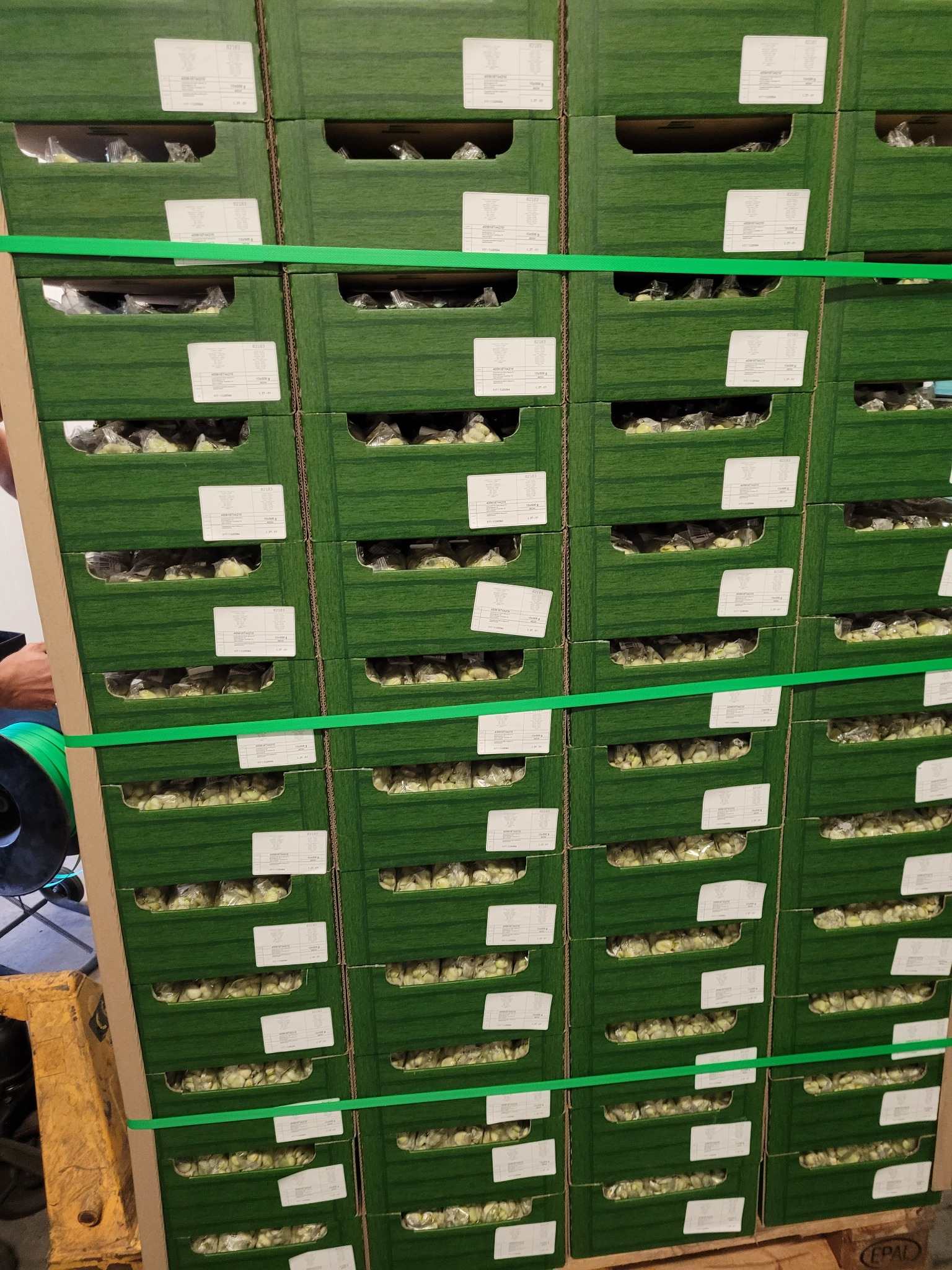 Paleta z zielonymi kartonami spiętymi taśmą, wypełnionymi zapakowanymi produktami spożywczymi. Widoczne etykiety na kartonach.