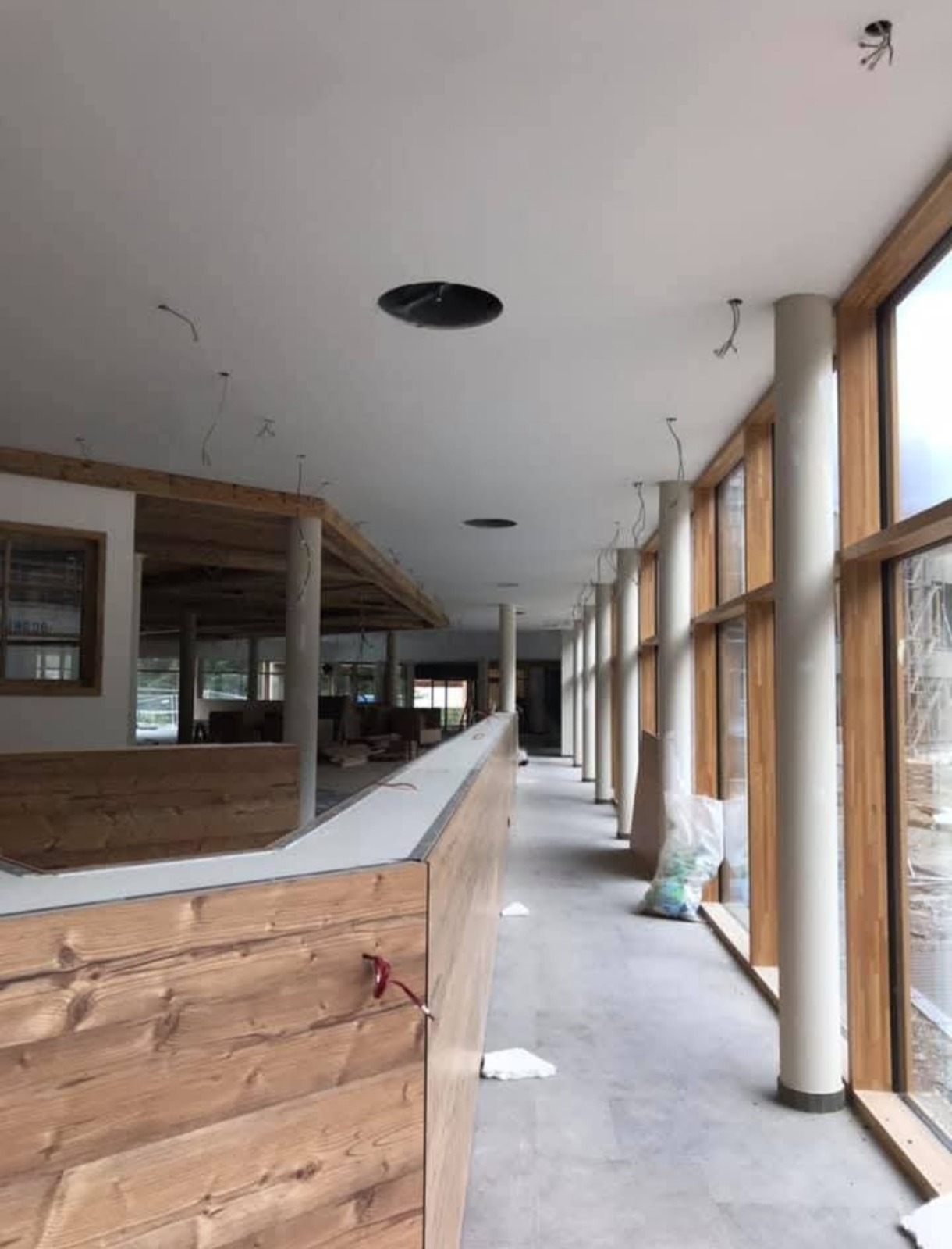 Wnętrze w trakcie wykańczania: drewniana zabudowa, kolumny i okna, widoczne instalacje w suficie. Szary beton na podłodze, brak wykończenia.