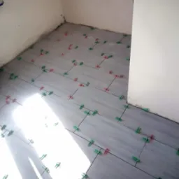 Układanie szarych płytek podłogowych z użyciem zielonych i czerwonych krzyżyków dystansowych w rogu pomieszczenia. Widoczne fugi i przygotowane podłoże.