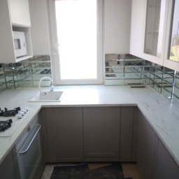 Agata Zając - Jasna kuchnia z marmurowym blatem i lustrzanymi płytkami na ścianie. Widoczne okno i zabudowa kuchenna. Nowoczesny design z elementami odbijającymi światło.