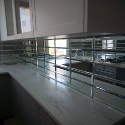 Agata Zając - Kuchnia po remoncie: lustrzane płytki nad blatem w szarym odcieniu, odbijające wnętrze. Widoczne białe szafki i sprzęty. Nietypowe wykończenie.