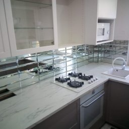 Agata Zając - Wykończona kuchnia z białymi szafkami, marmurowym blatem i lustrzaną mozaiką nad blatem. Widoczna płyta grzewcza i zlew. Nowoczesny design.