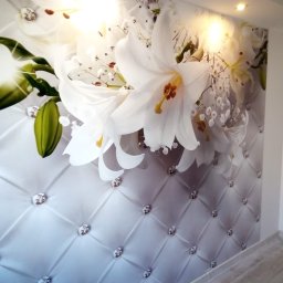 Agata Zając - Ściana z fototapetą: białe lilie na tle pikowanego materiału z kryształkami. Oświetlenie punktowe podkreśla wzór. Wykończenie wnętrza w jasnych barwach.