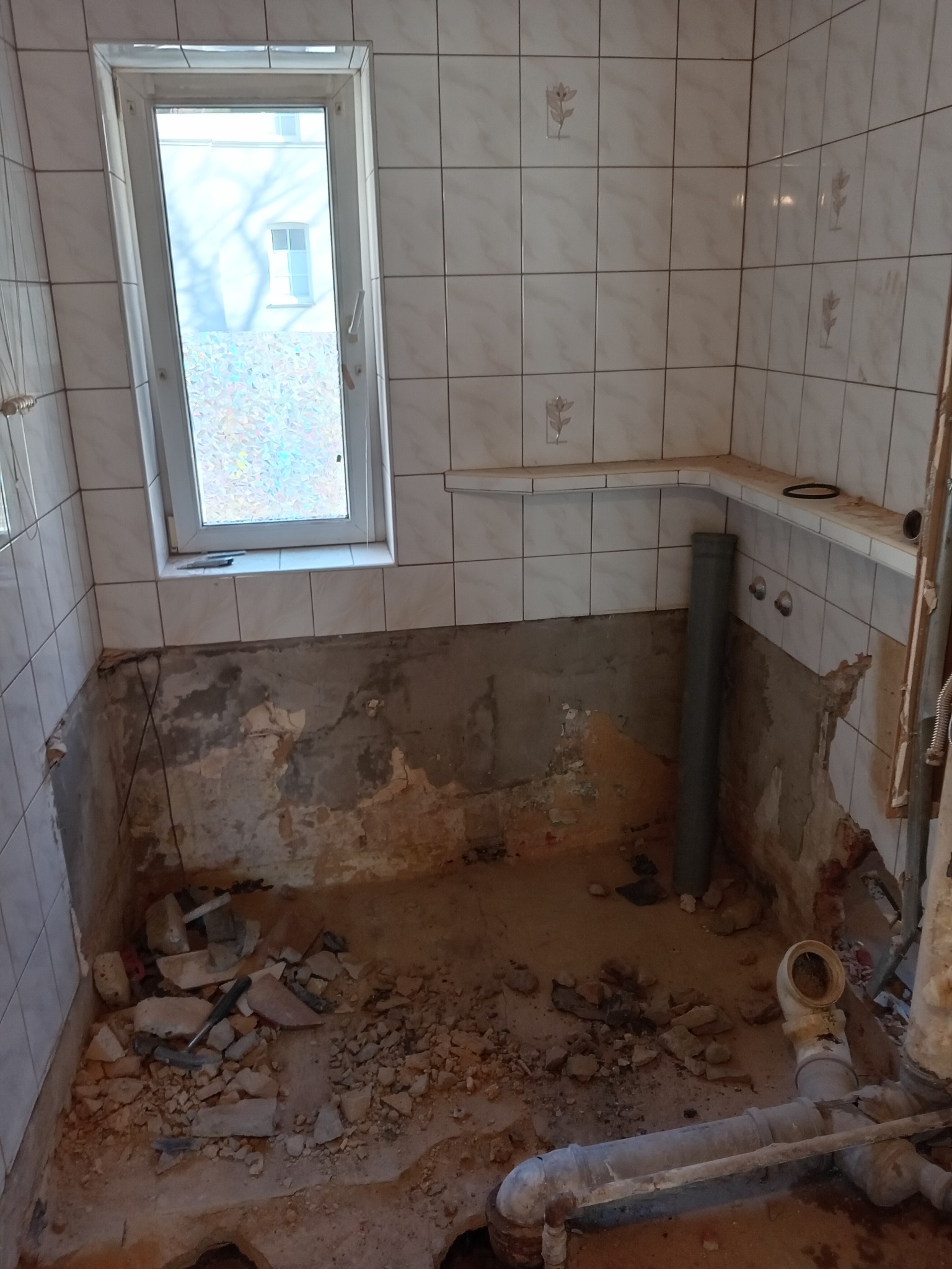 Łazienka w trakcie remontu: zerwane płytki, widoczne instalacje, okno z ozdobną folią. Etap przygotowania do dalszych prac wykończeniowych.