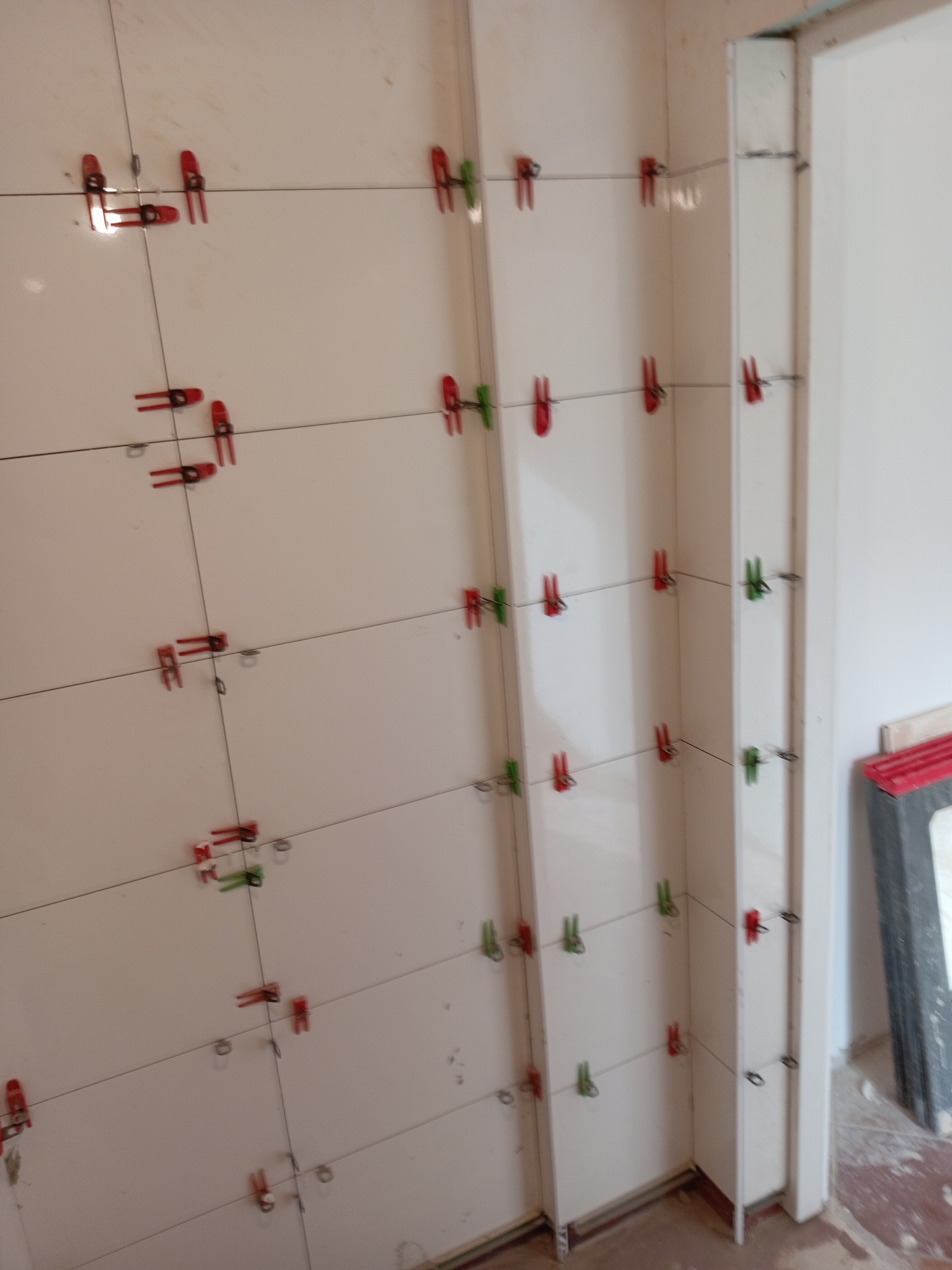 Ściana z białych płytek, zabezpieczonych czerwono-zielonymi klipsami poziomującymi, w trakcie prac wykończeniowych w łazience. Widoczny fragment futryny.