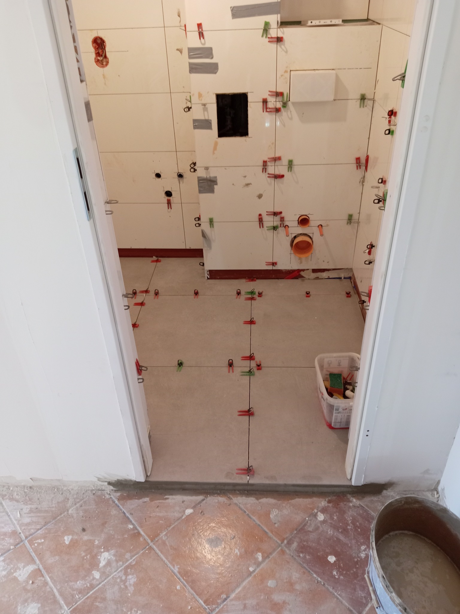 Łazienka w trakcie układania płytek: białe płytki na ścianach z elementami mocującymi, szare płytki na podłodze, widok zza progu.