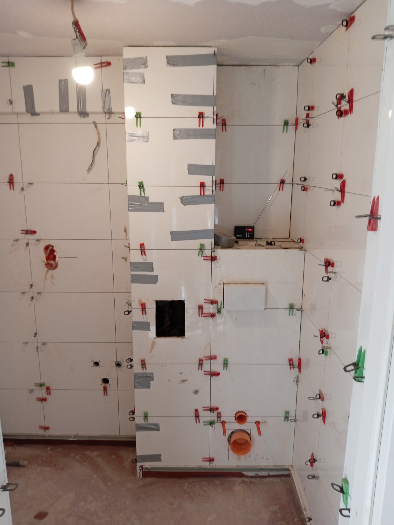 Ściany w trakcie prac glazurniczych: białe płytki, poziomowanie z użyciem klipsów i klinów, widoczne otwory na instalacje. Radio na półce. Podłoga nie wykończona.