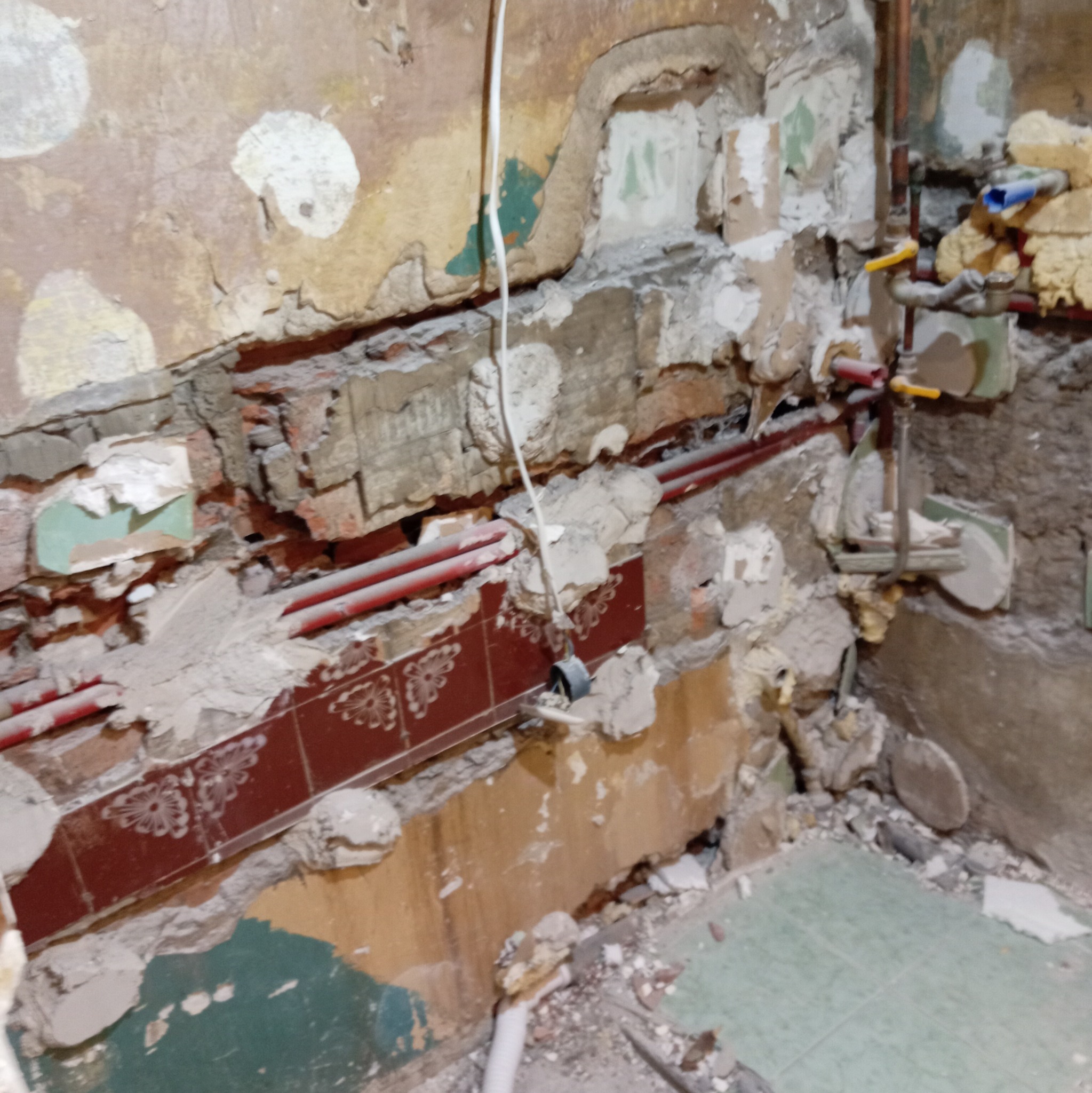 Demontaż starej instalacji w łazience. Odsłonięte rury, fragmenty płytek i gruzu na ścianach i podłodze. Widoczne pozostałości po remoncie.