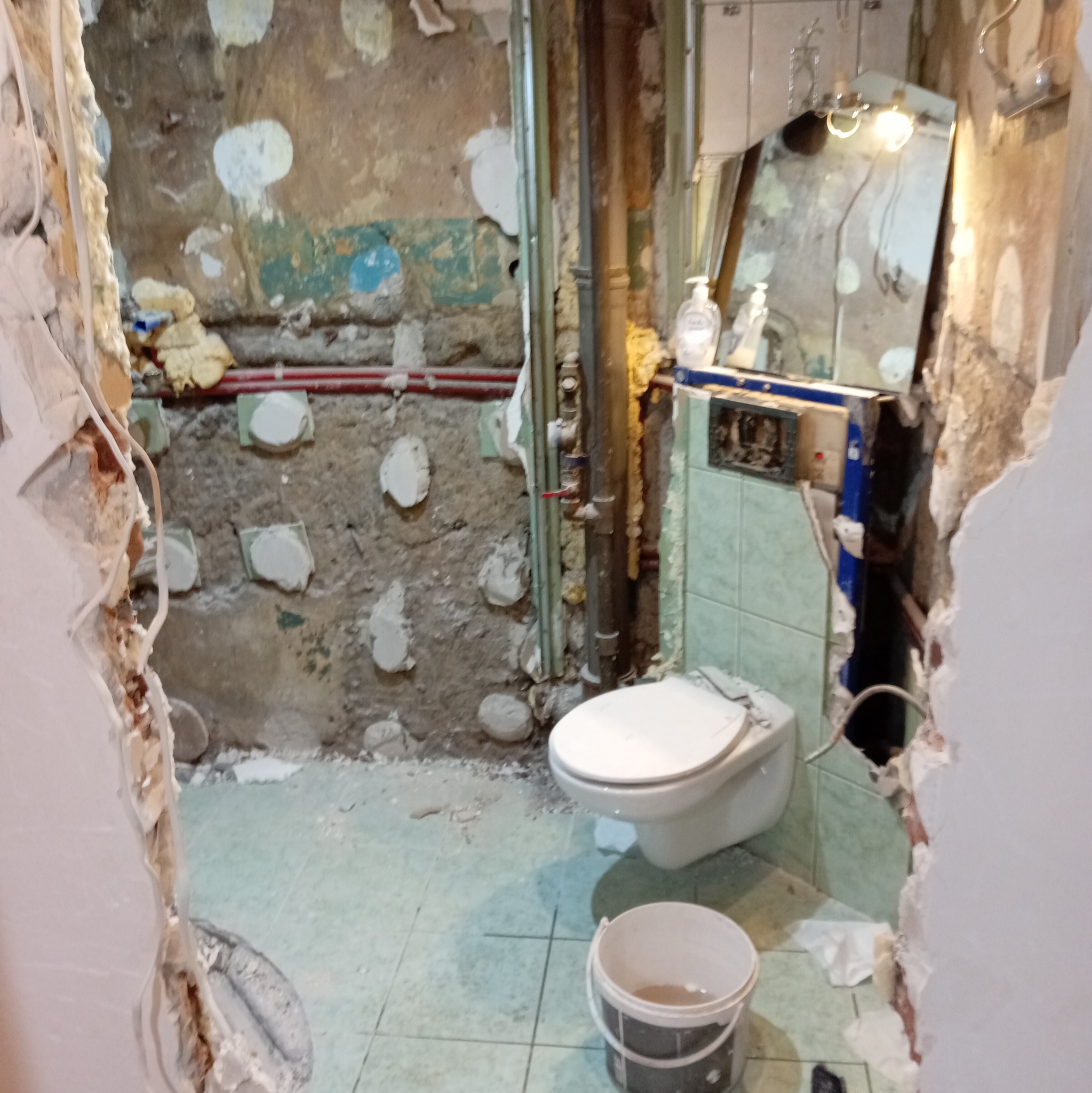 Łazienka w trakcie demontażu, z widocznymi rurami, fragmentami płytek i gruzem. Ujęcie z perspektywy wejścia, z wiadrem na podłodze.