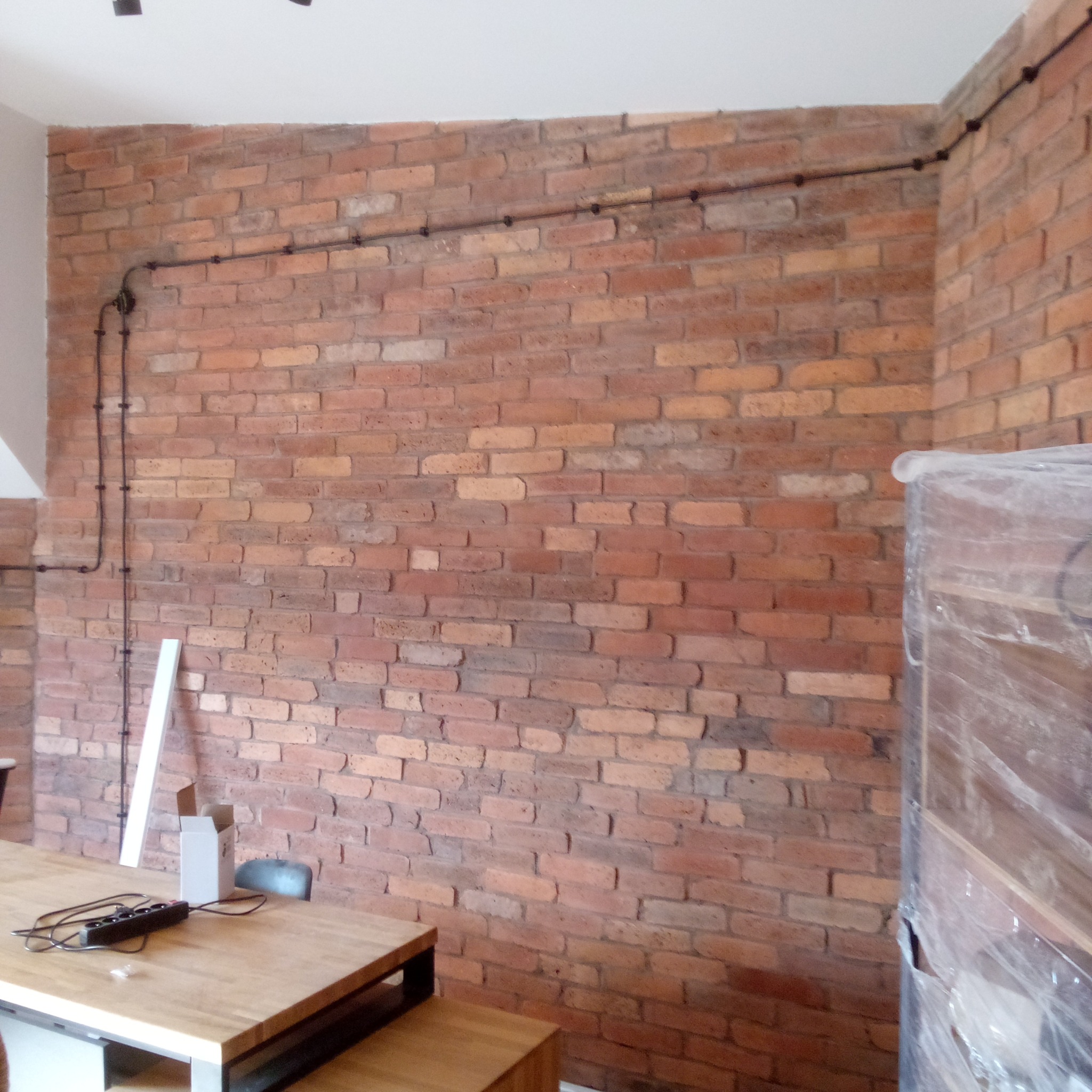 Ściana z czerwonej cegły z widoczną instalacją elektryczną. Na pierwszym planie stół z przedłużaczem. W tle częściowo widoczne elementy wyposażenia wnętrza.