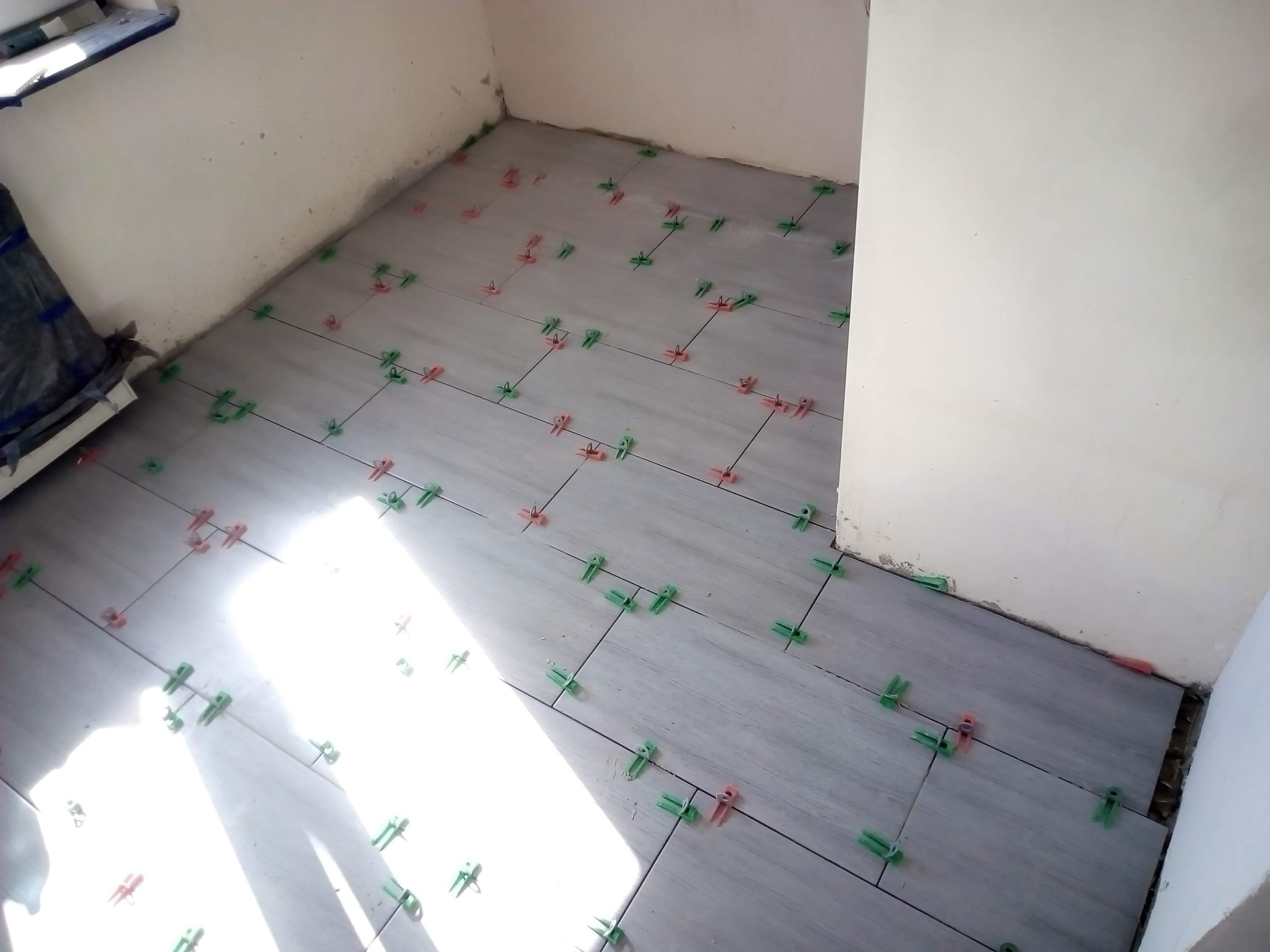 Układanie szarych płytek podłogowych z użyciem zielonych i czerwonych krzyżyków dystansowych w rogu pomieszczenia. Widoczne fugi i przygotowane podłoże.