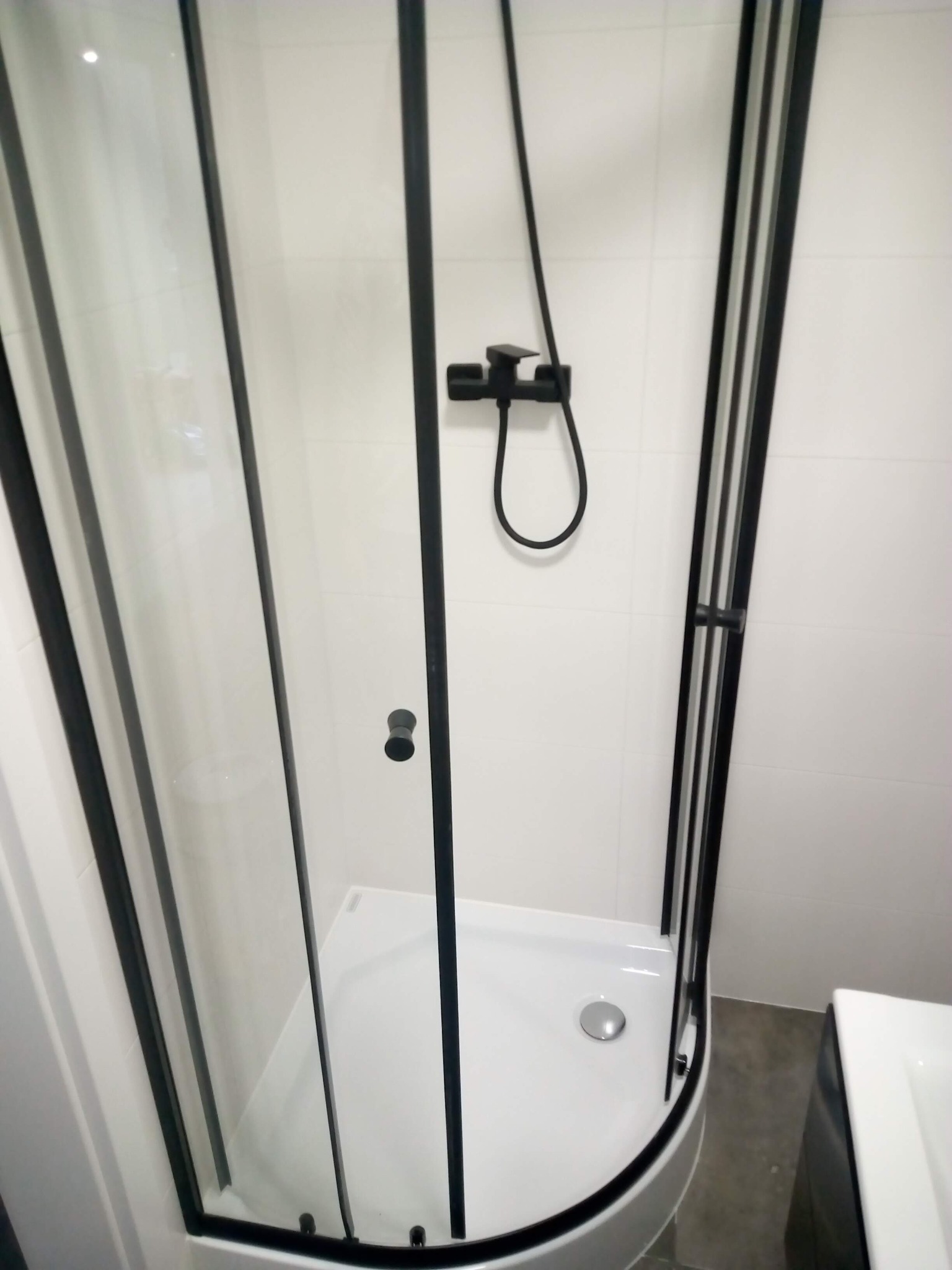 Nowoczesna kabina prysznicowa z czarnymi profilami i białą brodzikiem, na tle białych płytek. Minimalistyczny design, widoczny odpływ i bateria prysznicowa.