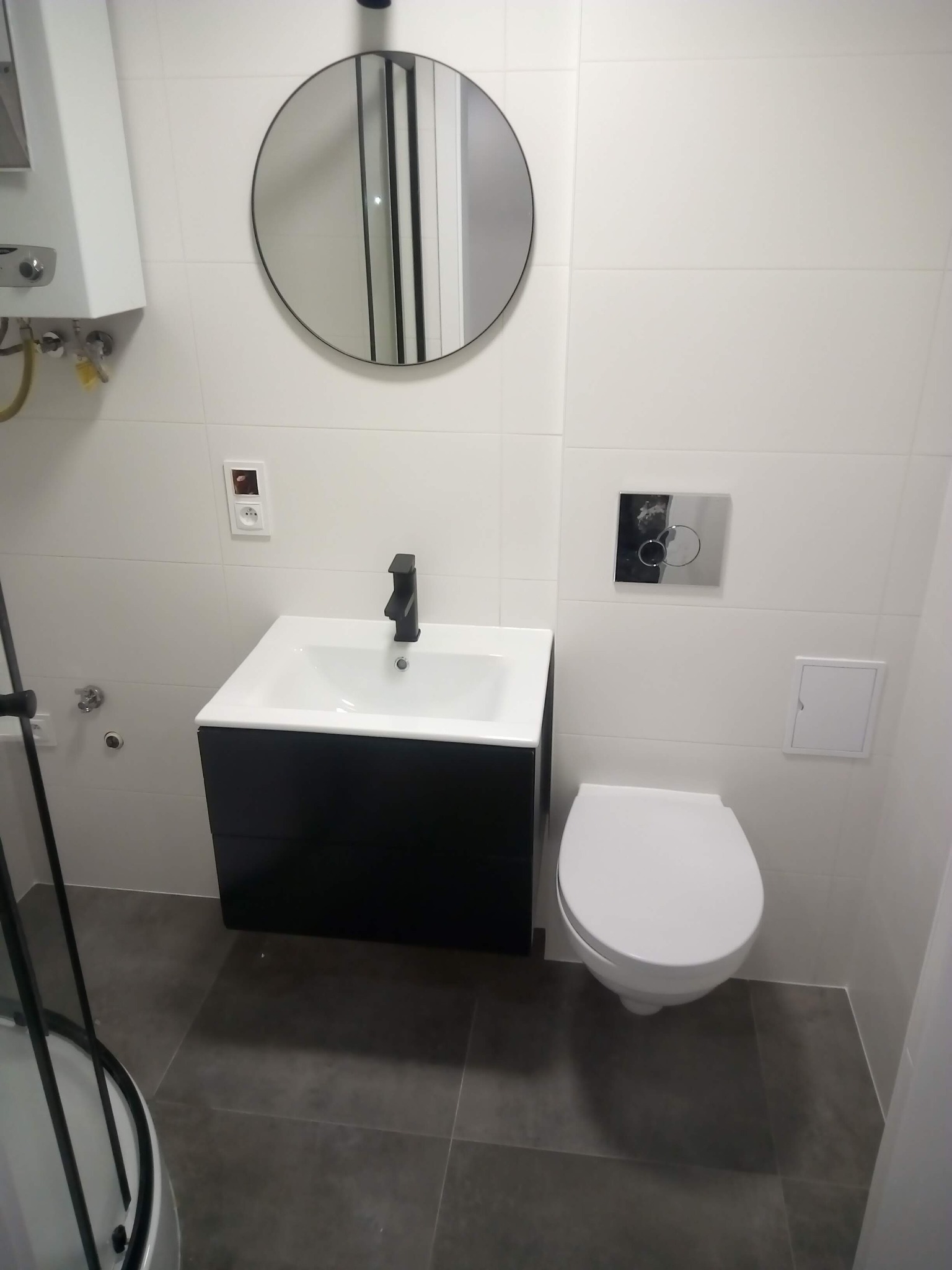 Nowoczesna łazienka z umywalką na szafce, wiszącą toaletą i okrągłym lustrem. Minimalistyczny design z białymi płytkami i szarą podłogą.