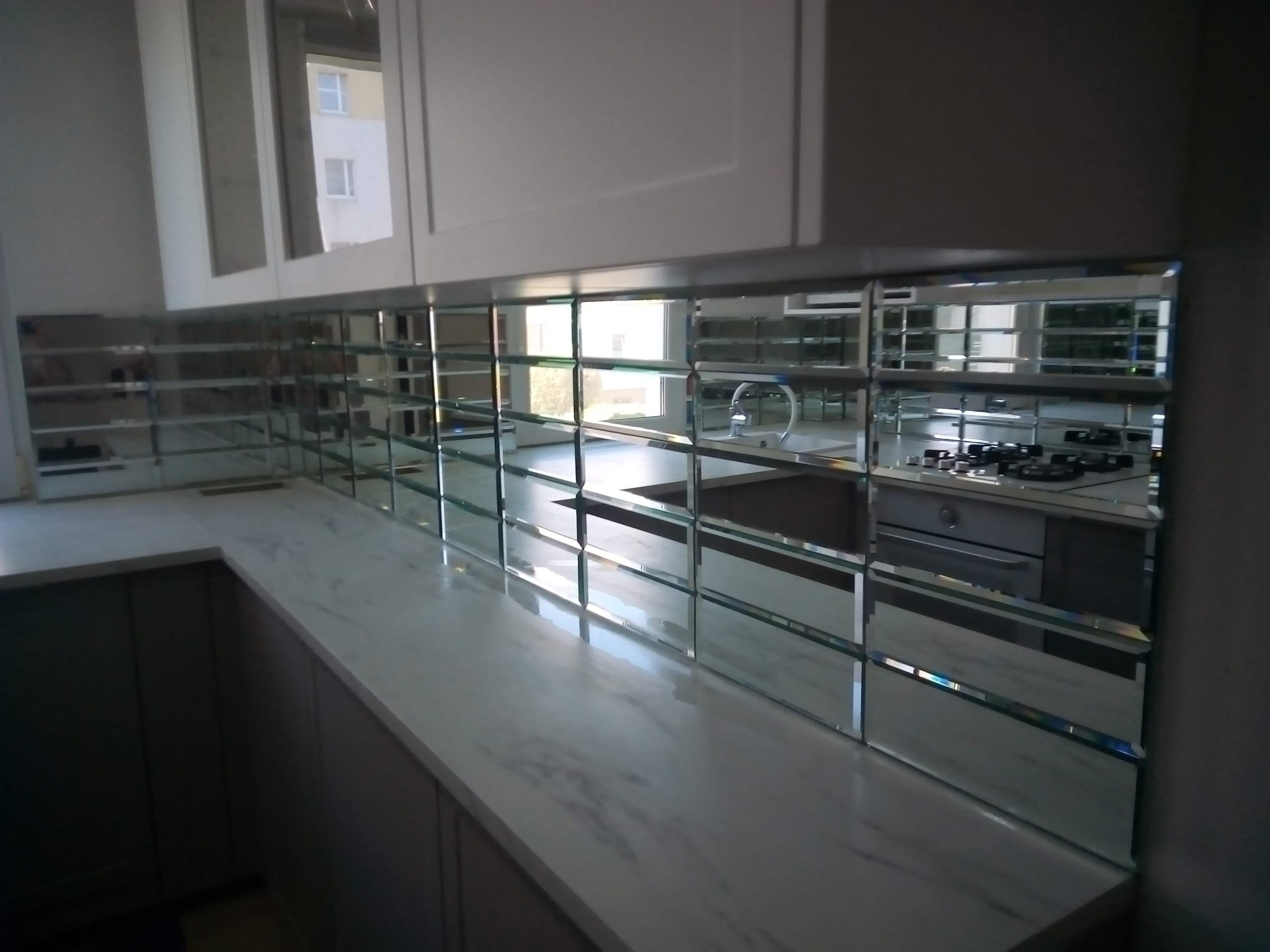 Kuchnia po remoncie: lustrzane płytki nad blatem w szarym odcieniu, odbijające wnętrze. Widoczne białe szafki i sprzęty. Nietypowe wykończenie.