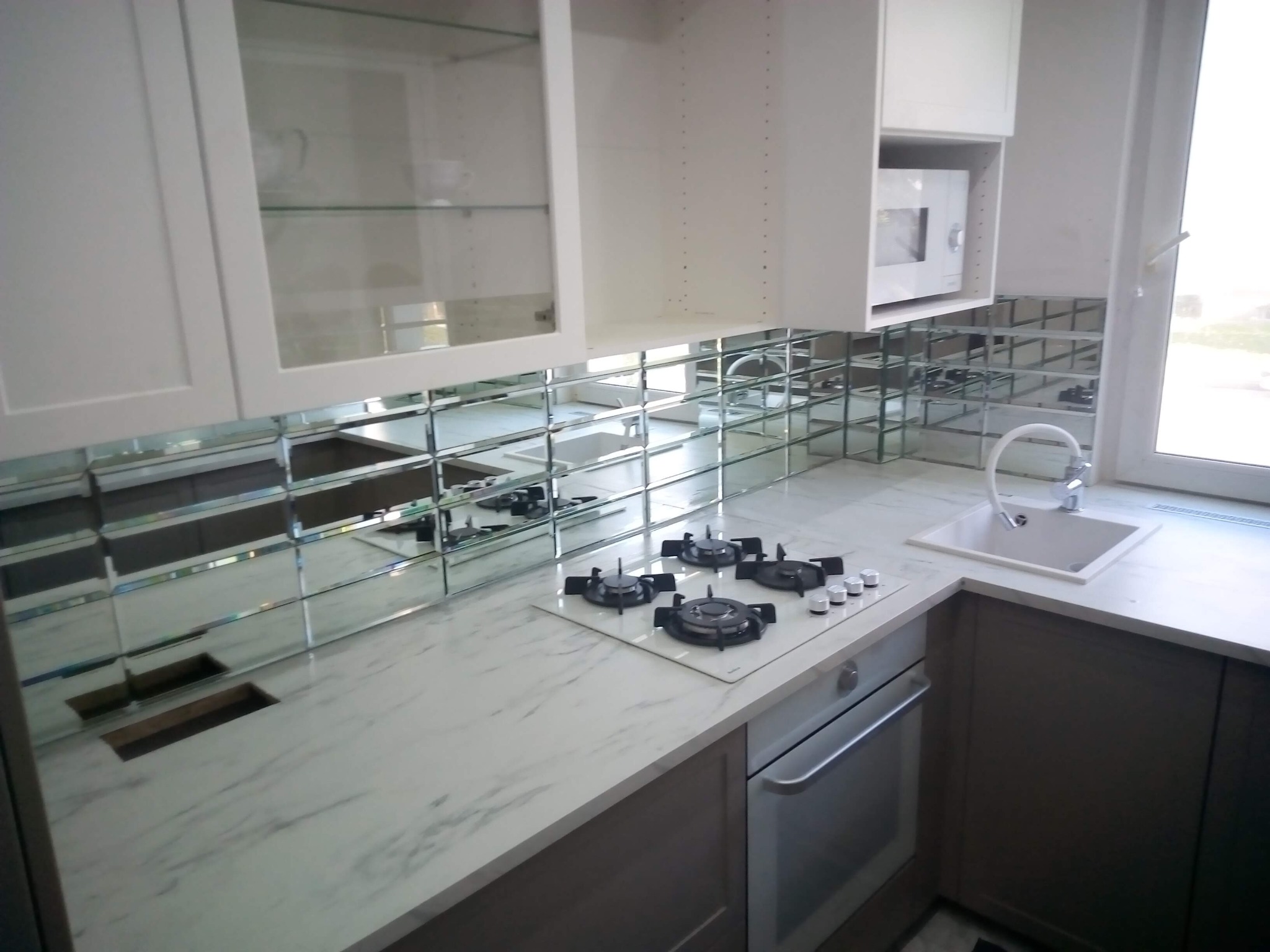 Wykończona kuchnia z białymi szafkami, marmurowym blatem i lustrzaną mozaiką nad blatem. Widoczna płyta grzewcza i zlew. Nowoczesny design.