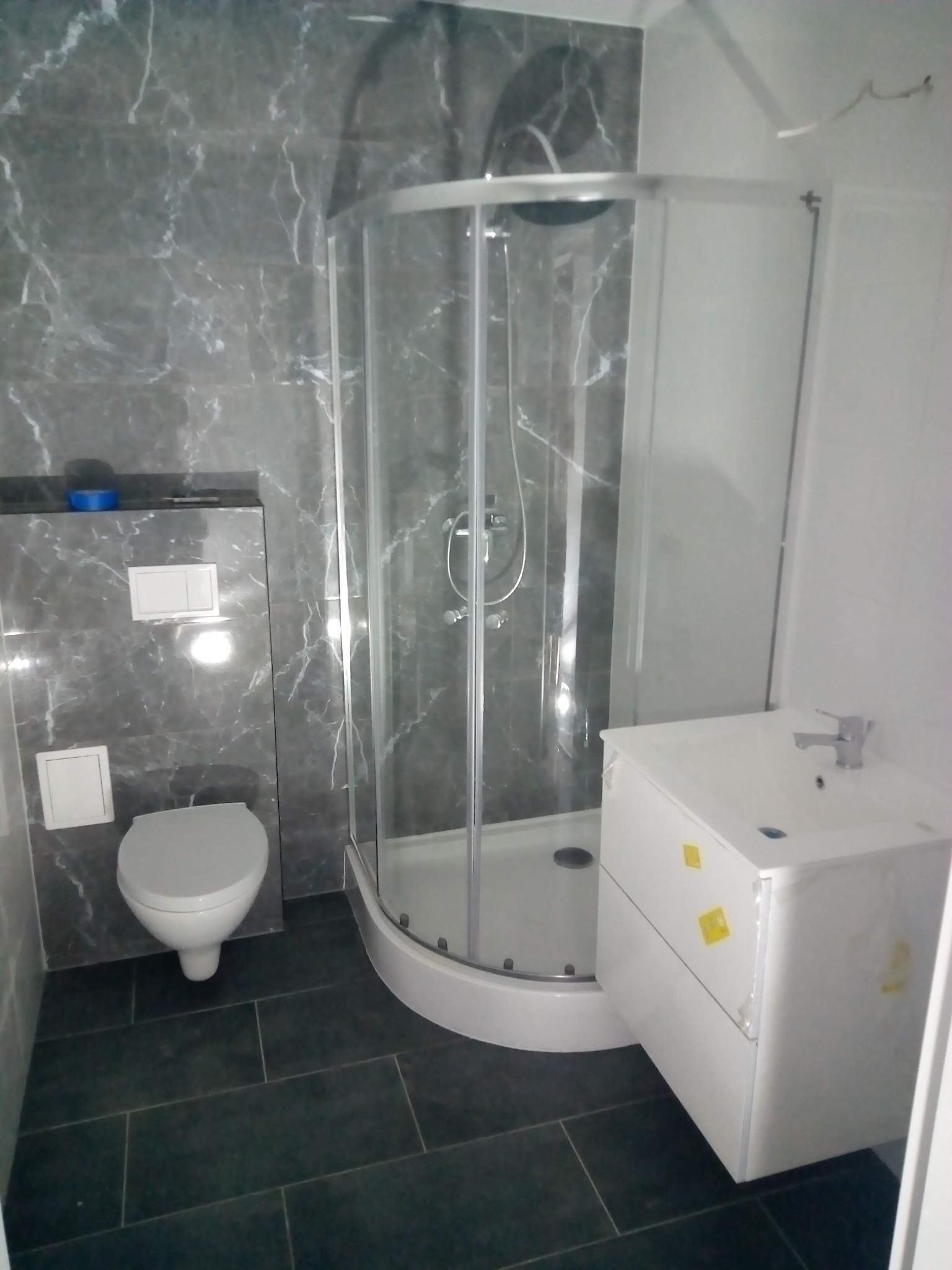 Wykończona łazienka z kabiną prysznicową, umywalką z szafką i toaletą w szaro-białej kolorystyce, płytki imitujące marmur na ścianach.