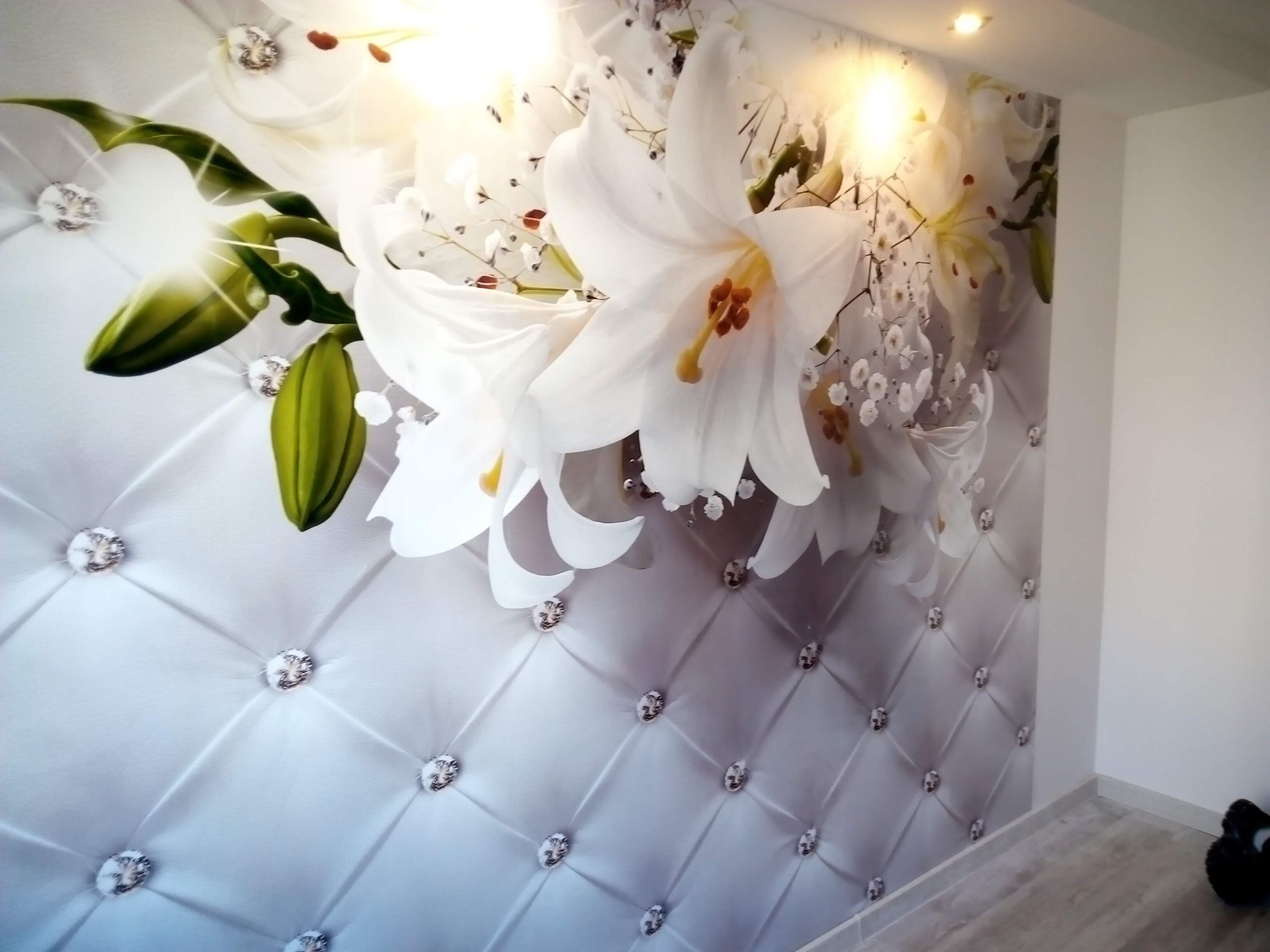 Ściana z fototapetą: białe lilie na tle pikowanego materiału z kryształkami. Oświetlenie punktowe podkreśla wzór. Wykończenie wnętrza w jasnych barwach.