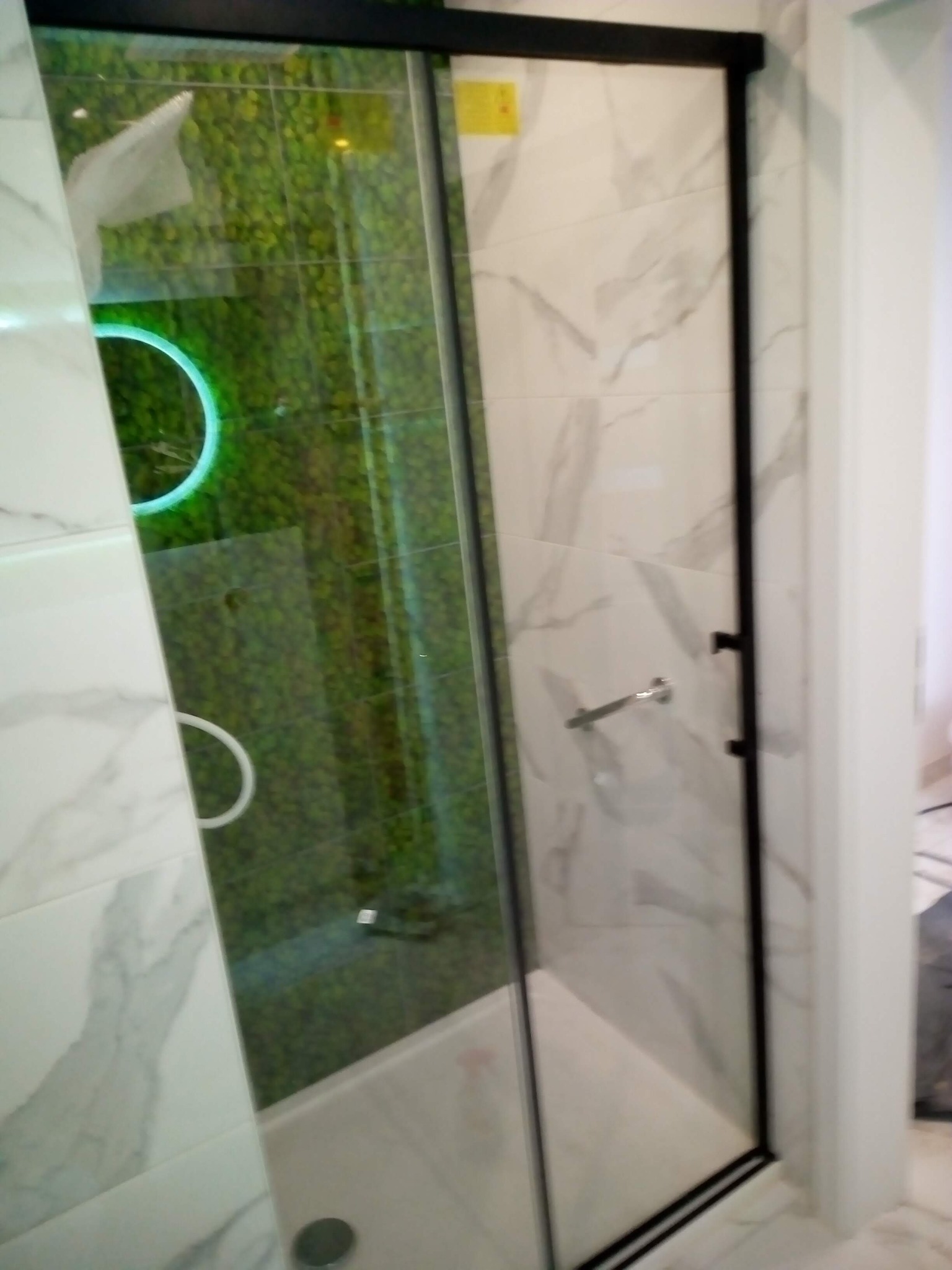 Nowoczesna kabina prysznicowa z przesuwnymi drzwiami, wykończona marmurowymi płytkami i podświetlanym panelem z zielenią. Czarna rama kabiny.