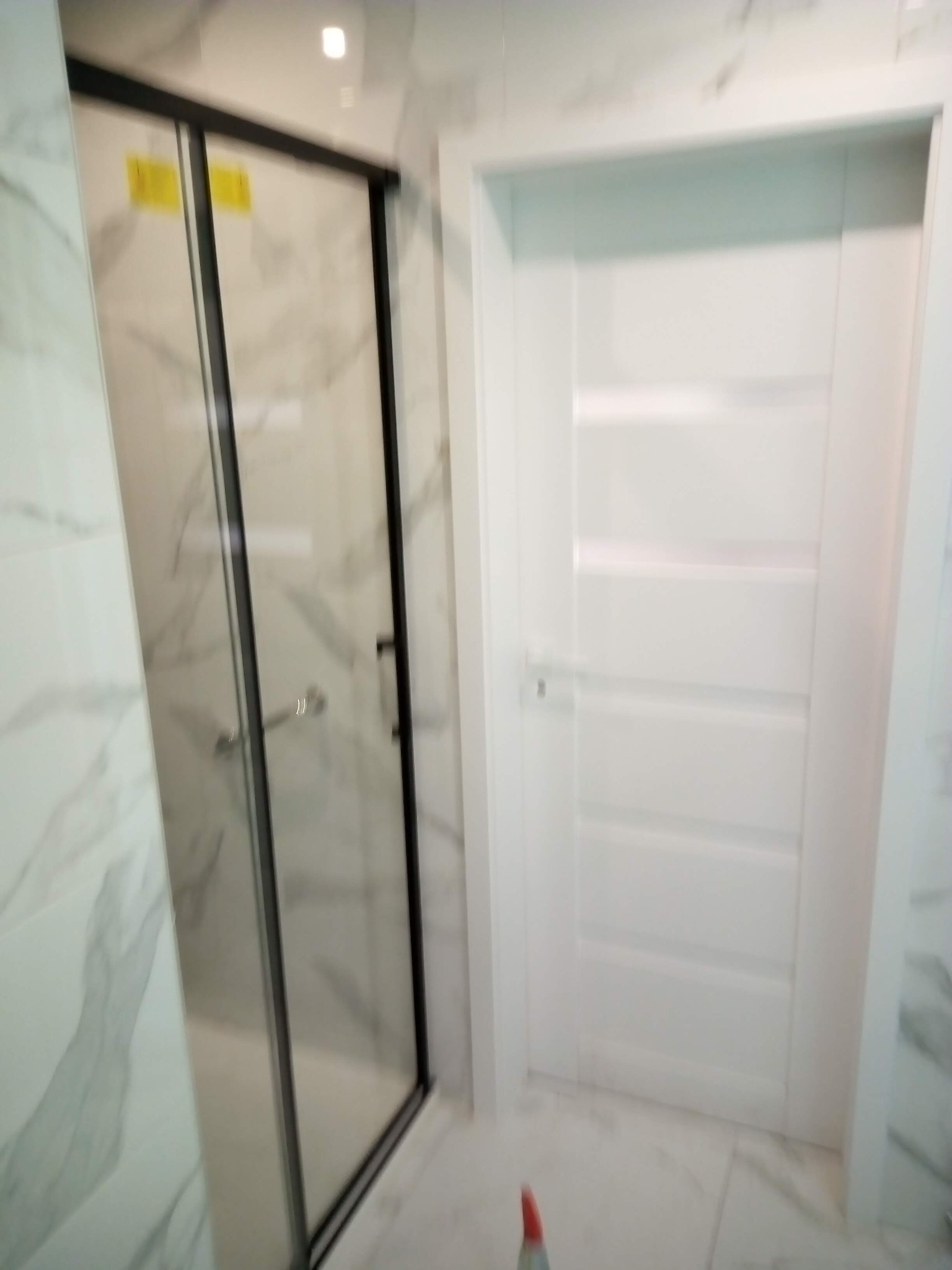 Nowoczesna łazienka z kabiną prysznicową typu walk-in z czarnymi profilami i białą, panelową ścianą z drzwiami. Marmurowe płytki na ścianach i podłodze.