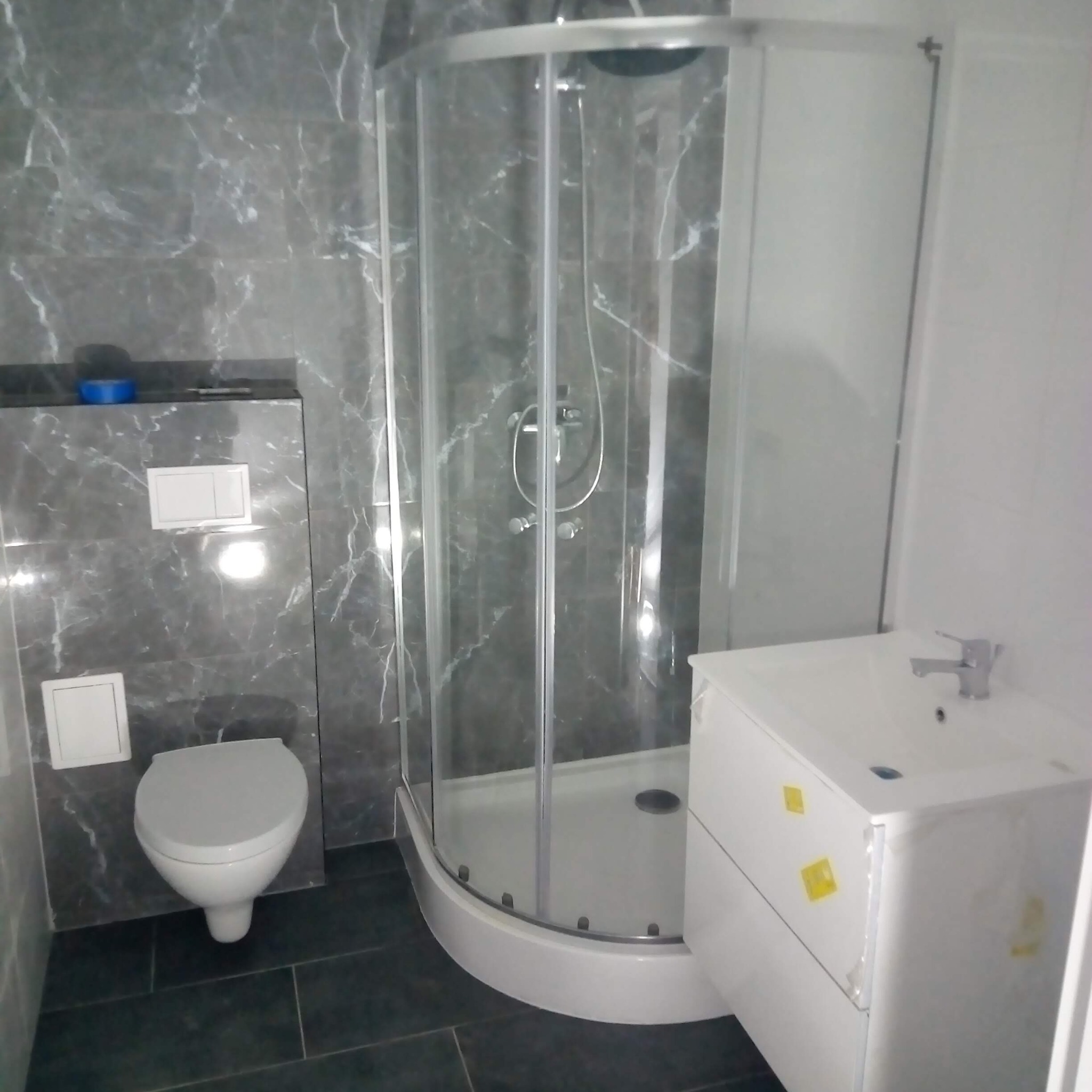 Wykończona łazienka: kabina prysznicowa, umywalka z szafką, toaleta podwieszana na tle szarych płytek imitujących marmur i ciemnej podłogi.