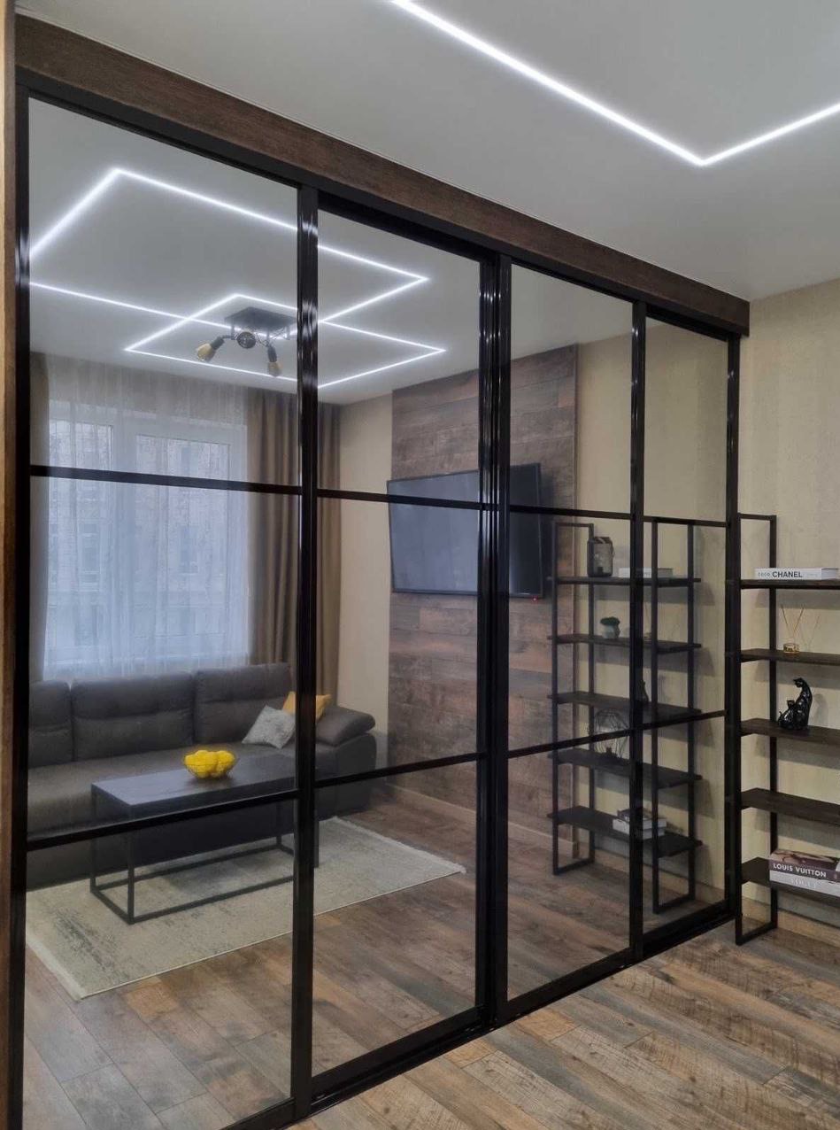 Nowoczesne drzwi przesuwne z czarnymi szprosami oddzielają salon. Widoczne oświetlenie sufitowe LED i regały. Minimalistyczny design, ciepłe odcienie drewna.