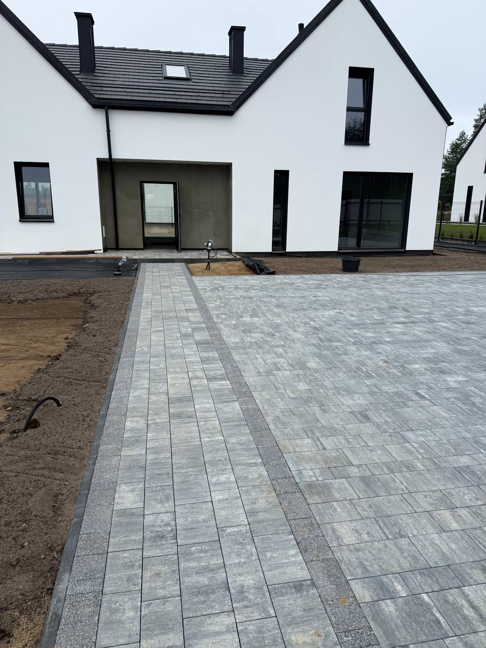 Nowoczesny podjazd z szarej kostki brukowej przed białym domem z czarnymi oknami i dachem. Widoczny fragment ogrodu i przygotowane podłoże pod dalsze prace.