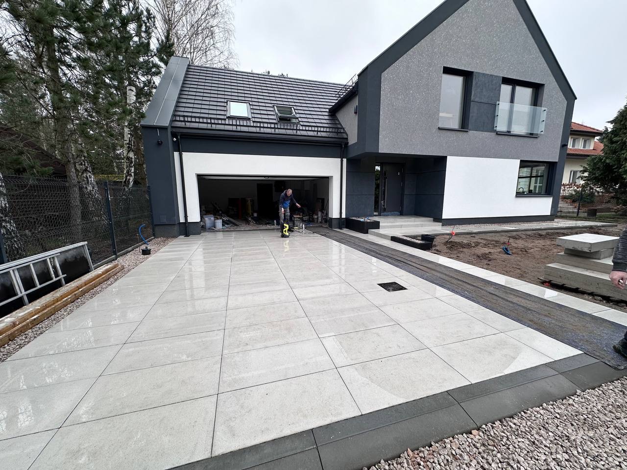 Nowoczesny podjazd z jasnych, kwadratowych płyt, obramowany ciemnoszarą kostką. W tle dom z garażem i pracownik używający myjki ciśnieniowej. Widoczne przygotowania do wykończenia.