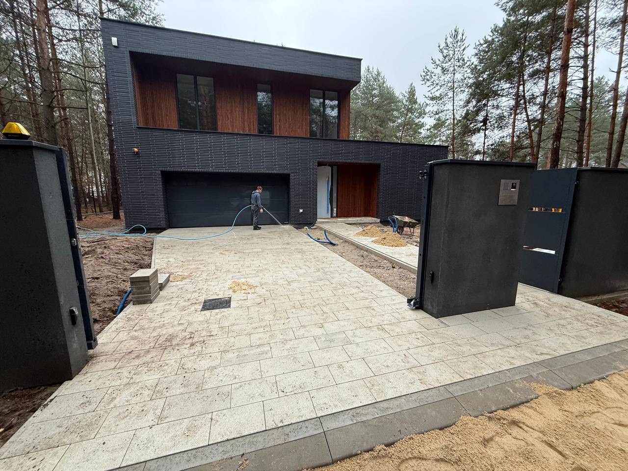 Nowoczesny dom z jasną kostką brukową, którą myje pracownik. Widoczne otwarte, grafitowe wrota wjazdowe i fragment elewacji z drewnianymi elementami.