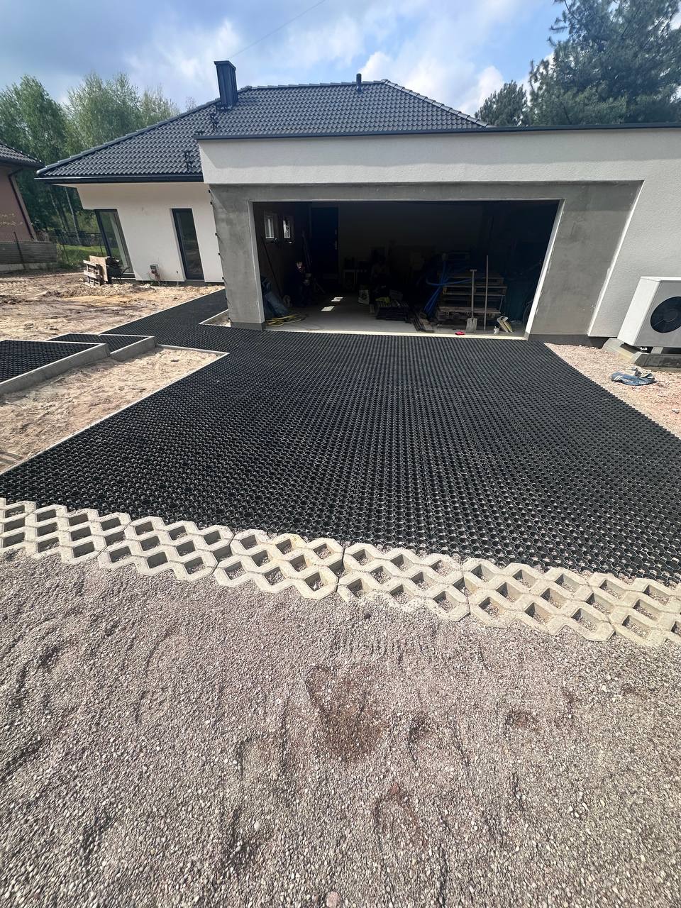 Częściowo ułożona, czarna, ażurowa nawierzchnia na podjeździe z obrzeżami z betonowych elementów. W tle dom z garażem i zielenią.