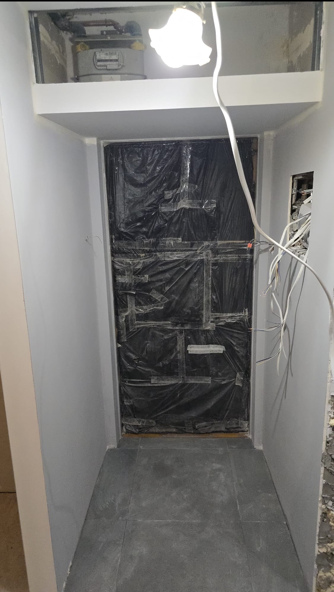 Korytarz w trakcie remontu: ściany pomalowane na szaro, podłoga wyłożona płytkami. Otwór zabezpieczony czarną folią i taśmą. Widoczne przewody elektryczne.